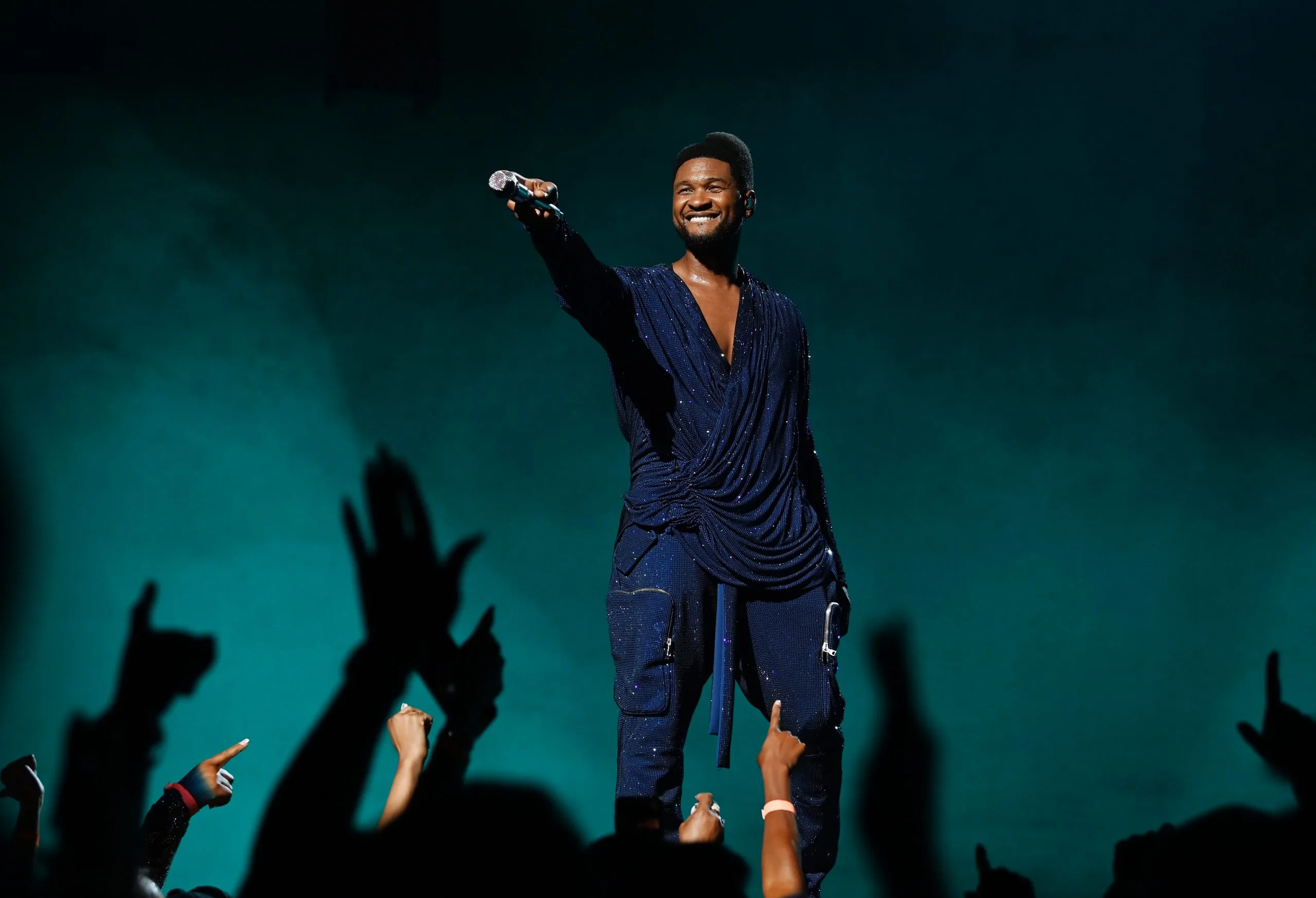 A photo of Usher performing at the Super Bowl LVIII halftime show, surrounded by a crowd of energetic dancers and illuminated by colorful stage lights.