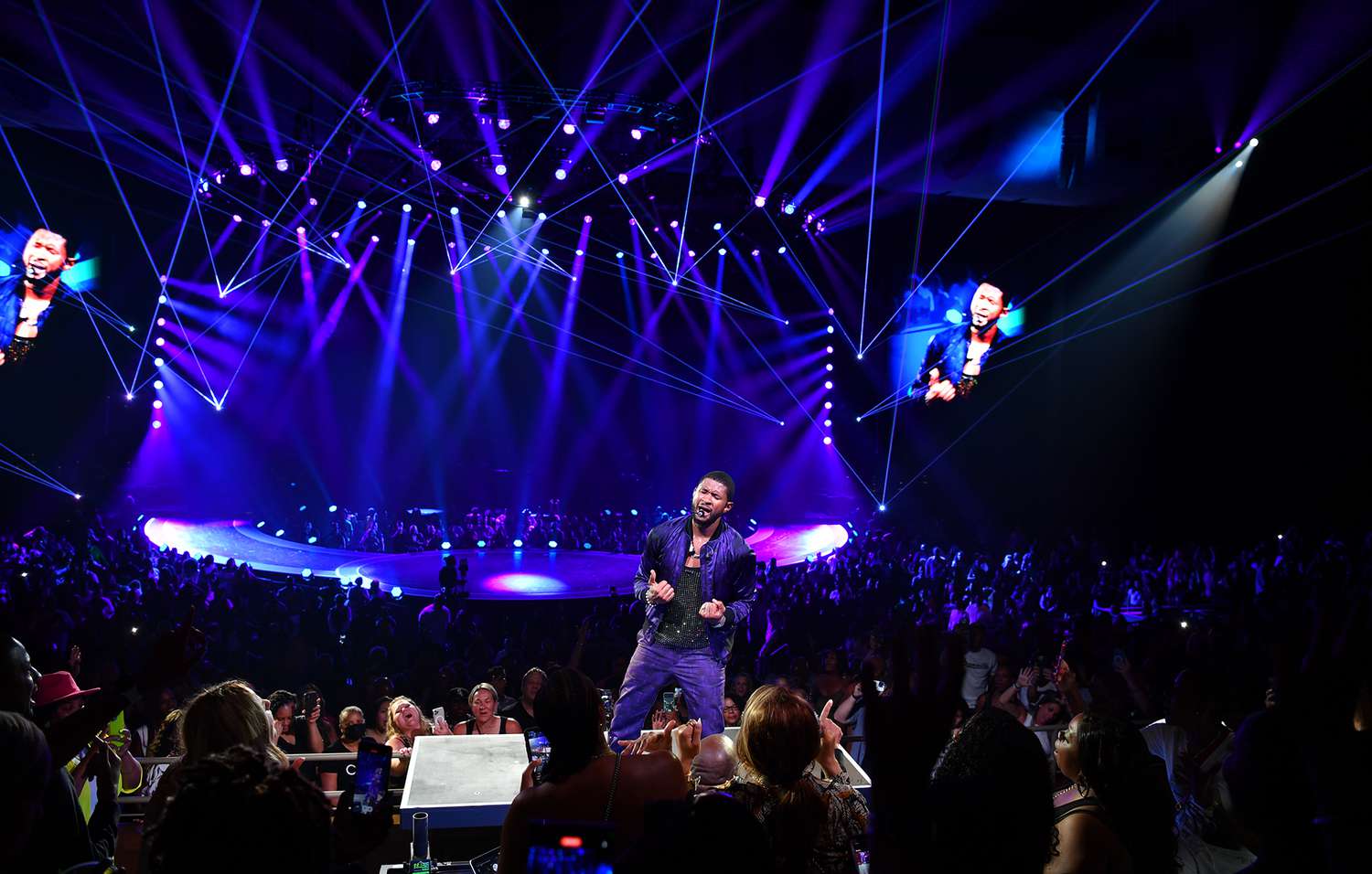 A photo of Usher performing at the Super Bowl LVIII halftime show, surrounded by a crowd of energetic dancers and illuminated by colorful stage lights.